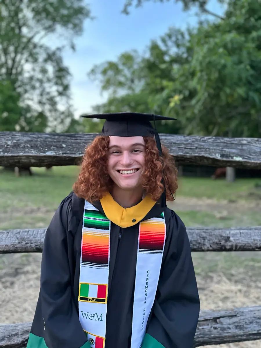 Isaiah in a William & Mary graduation cap and gown with stoles, smiling in front of a wooden fence